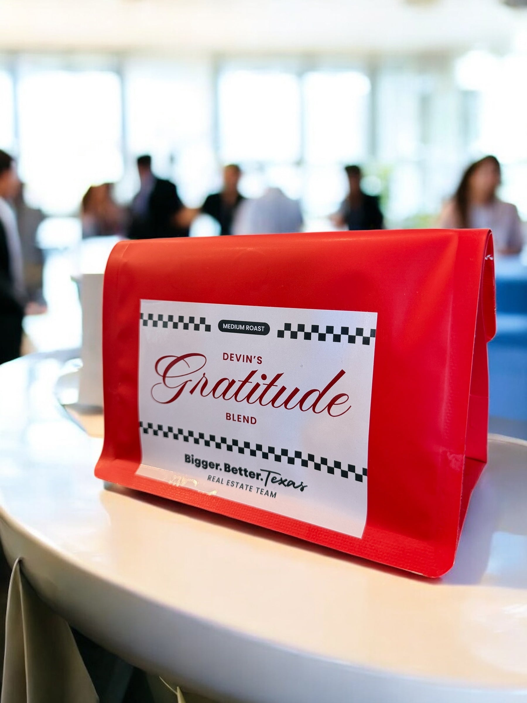 Red coffee bag labeled 'Devin's Gratitude Blend' on a table with blurred people in the background